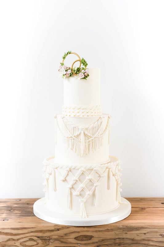 Three tier ivory boho wedding cake dressed with sugar macrame lace, topped with decorated gold rings by Saddleworth based Union Cakes