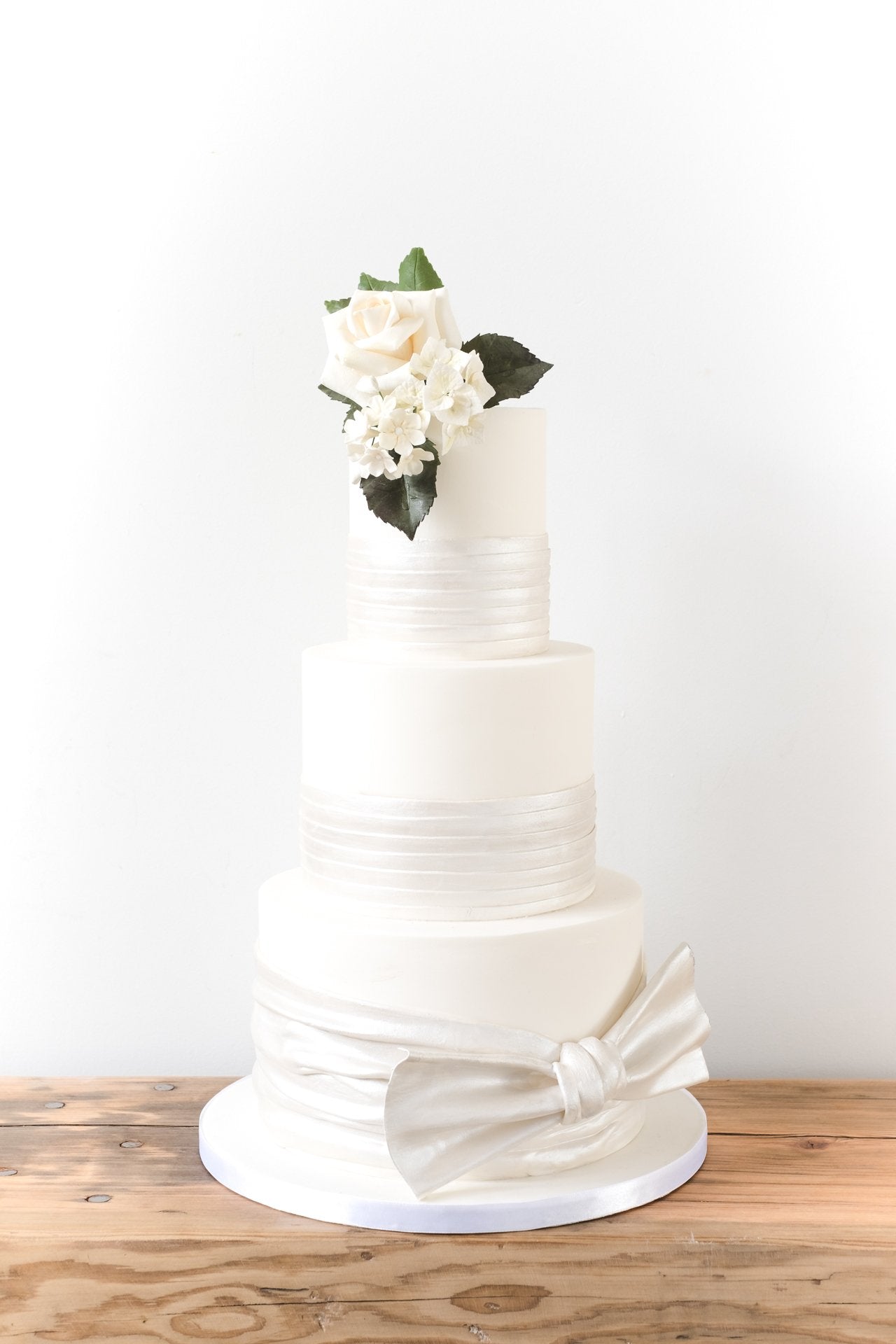 Three tier ivory wedding cake partially wrapped with pearlescent satin effect sugarpaste, topped with a white sugar rose
